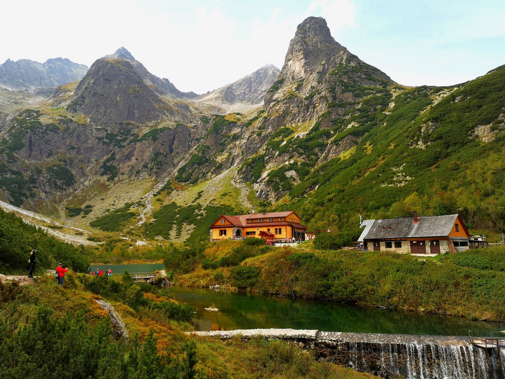 Charming mountain lodge by a lake, surrounded by dramatic peaks and greenery.