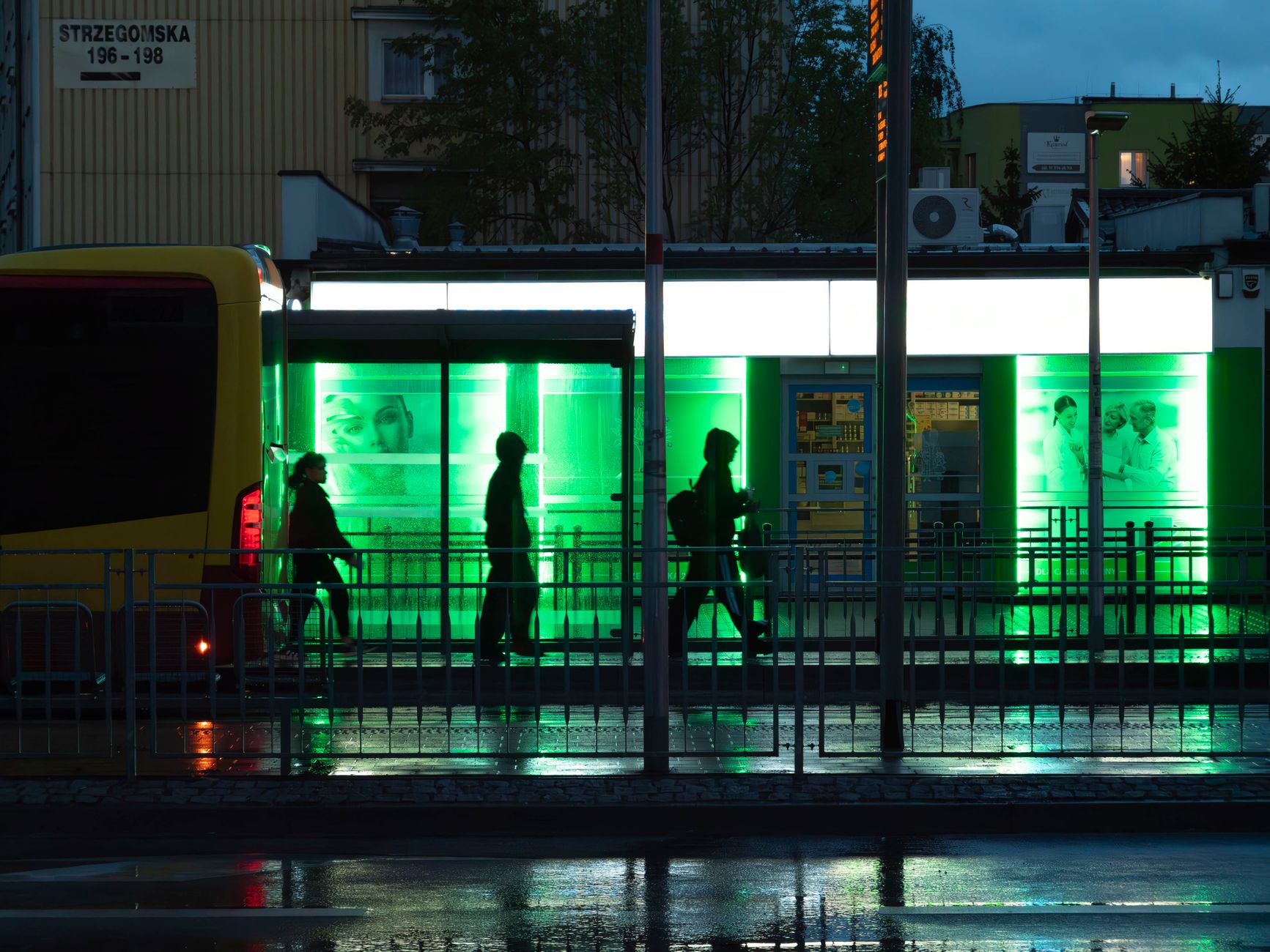 Silhouetted people at a bus stop on a rainy evening in Wrocław, Poland, with glowing green lights.