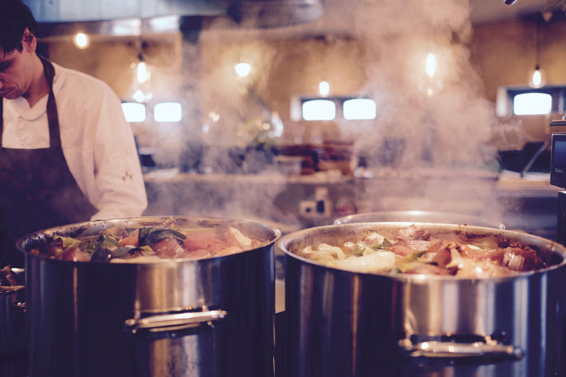 A chef in a restaurant kitchen tends to large pots of steaming soup, creating a cozy atmosphere.