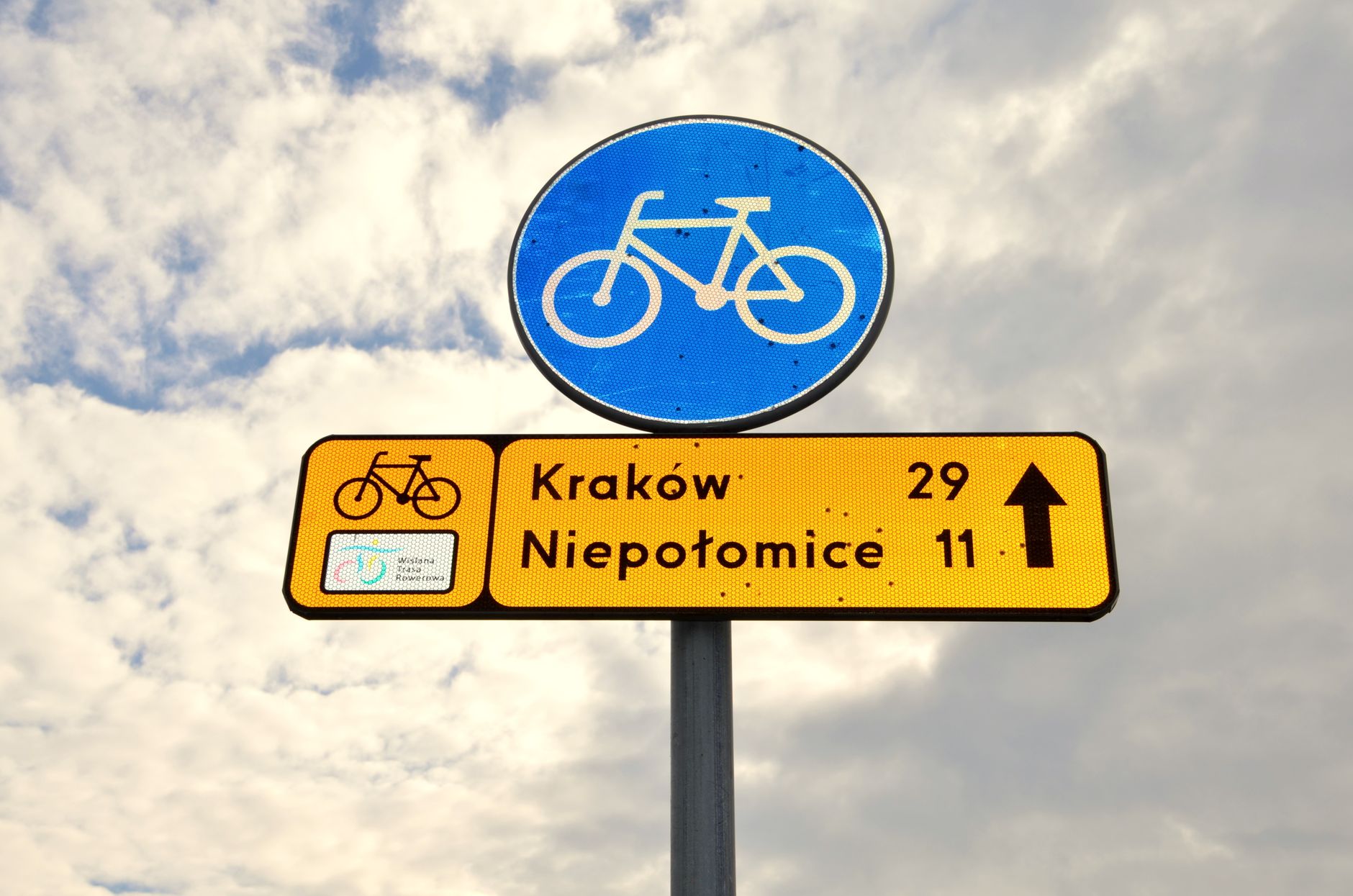 A bicycle route signpost indicating directions to Kraków and Niepołomice under a partly cloudy sky.