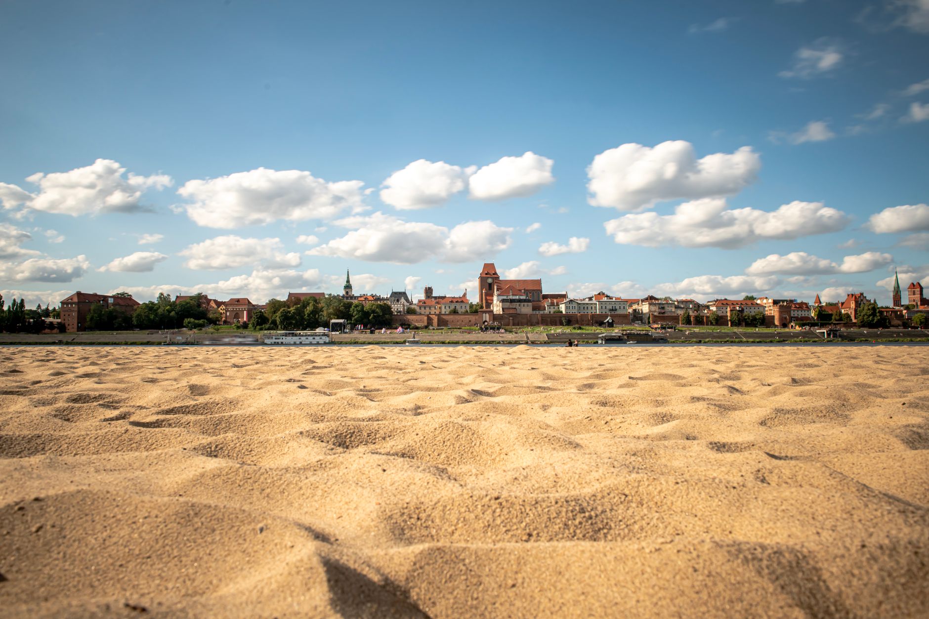 A scenic view of a sandy beach with a historic city skyline under a bright blue sky with clouds.