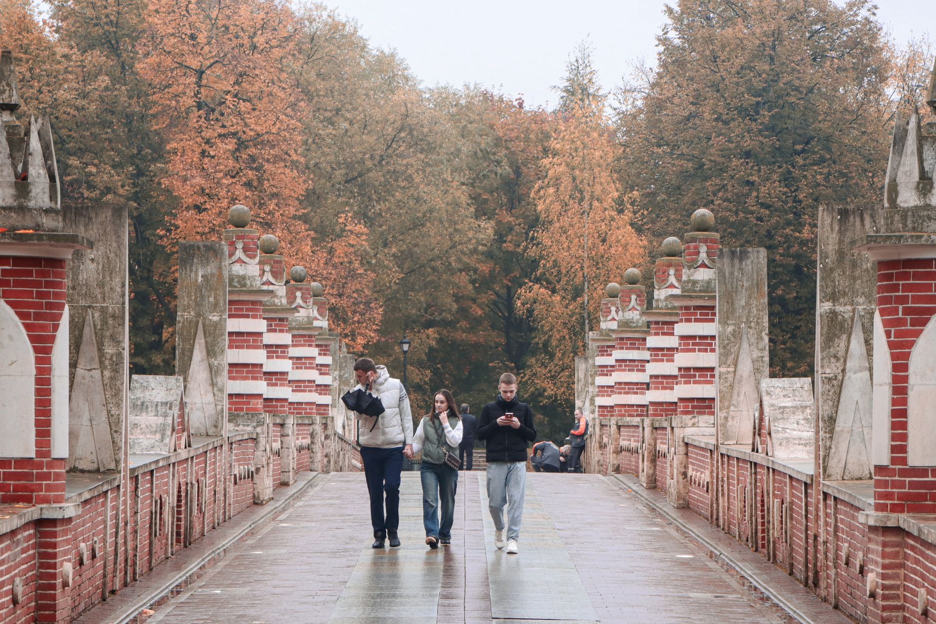 A group of friends enjoying a serene autumn walk across a historic red brick bridge surrounded by colorful foliage.