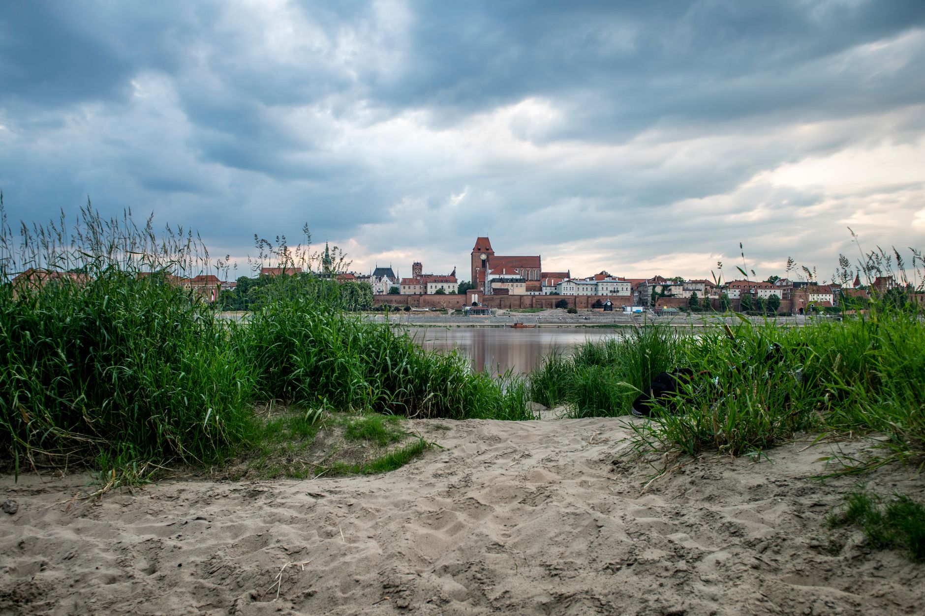 Cloudy sky over the historic Torun skyline viewed from a sandy riverbank.