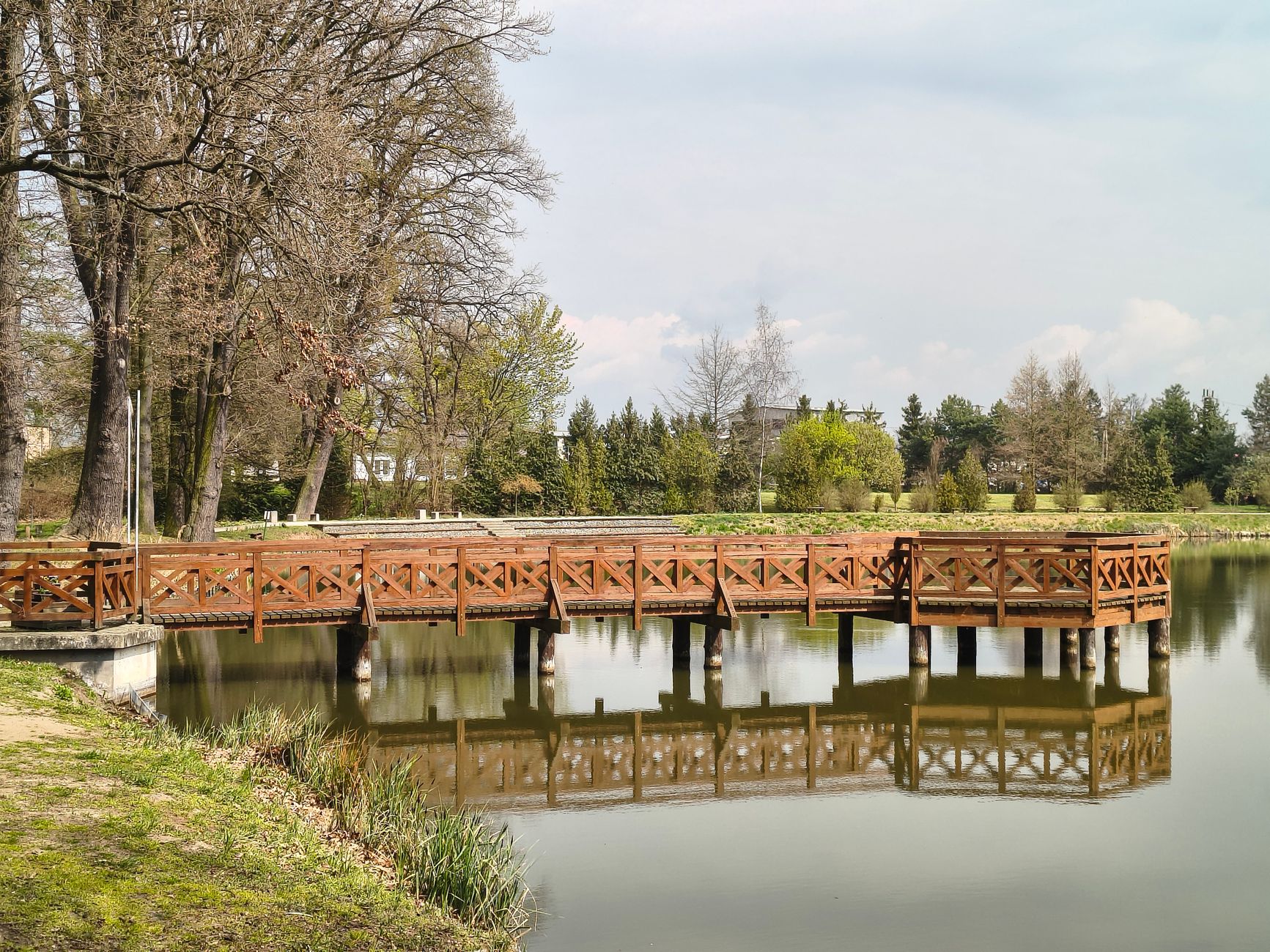 A serene view of a wooden pier reflecting in a calm pond surrounded by trees in a spring landscape.