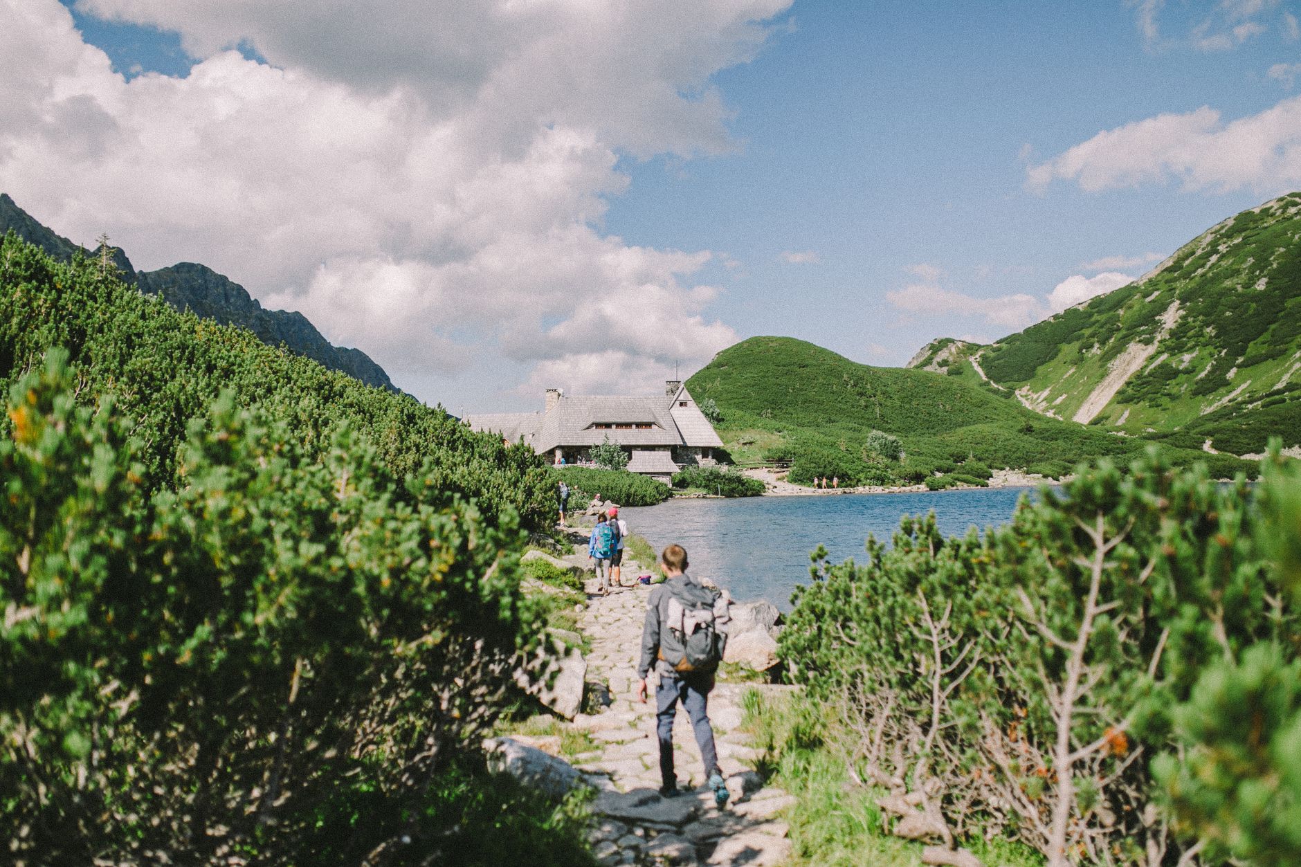 Hikers explore a picturesque mountain path by a serene lake under a vibrant sky.