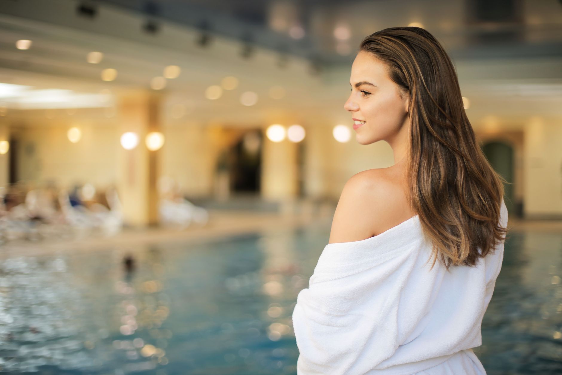 Portrait of a smiling woman in a robe by an indoor swimming pool, exuding relaxation and luxury.